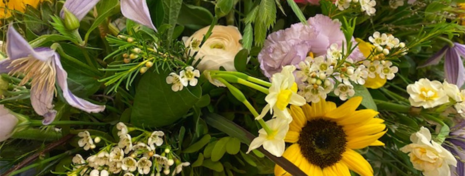 En farverig buket blomster med forskellige arter og farver, herunder solsikker, roser, og liljer, samt grønne blade. Blomsterne er tæt samlet og skaber et frodigt og livligt udtryk.