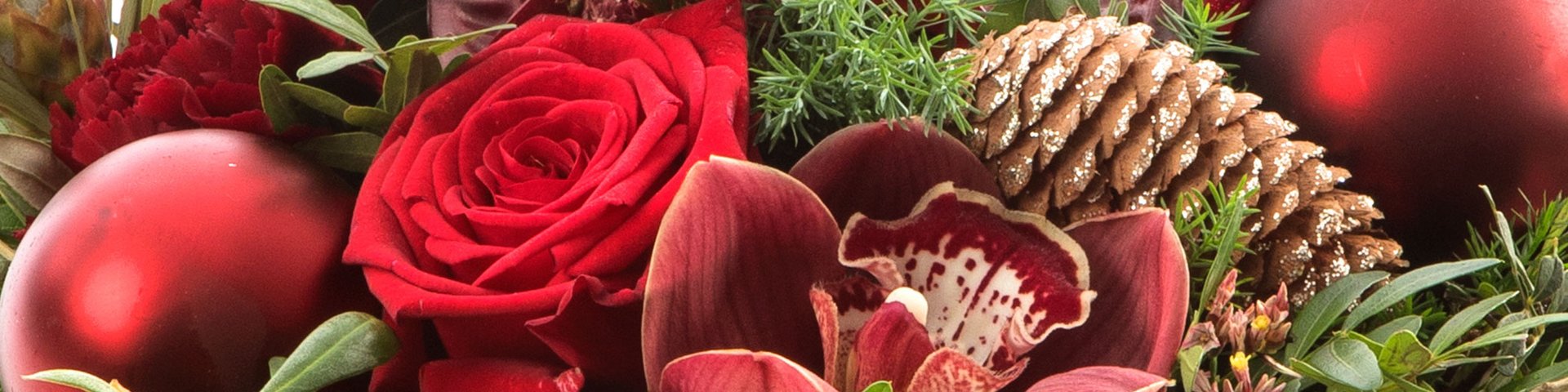 Blomsterarrangement med røde roser, en lyserød orkide og grønne blade, samt dekorationselementer som julepynt i rødt og en fyrrekogle. Det skaber en varm og festlig atmosfære, der indfanger essensen af julen.